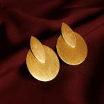 A pair of handcrafted gold-plated earrings featuring a matte brushed finish. The design includes a teardrop-shaped leaf layered over a circular disc, displayed on deep maroon satin fabric. Modern, minimalist, and luxurious.