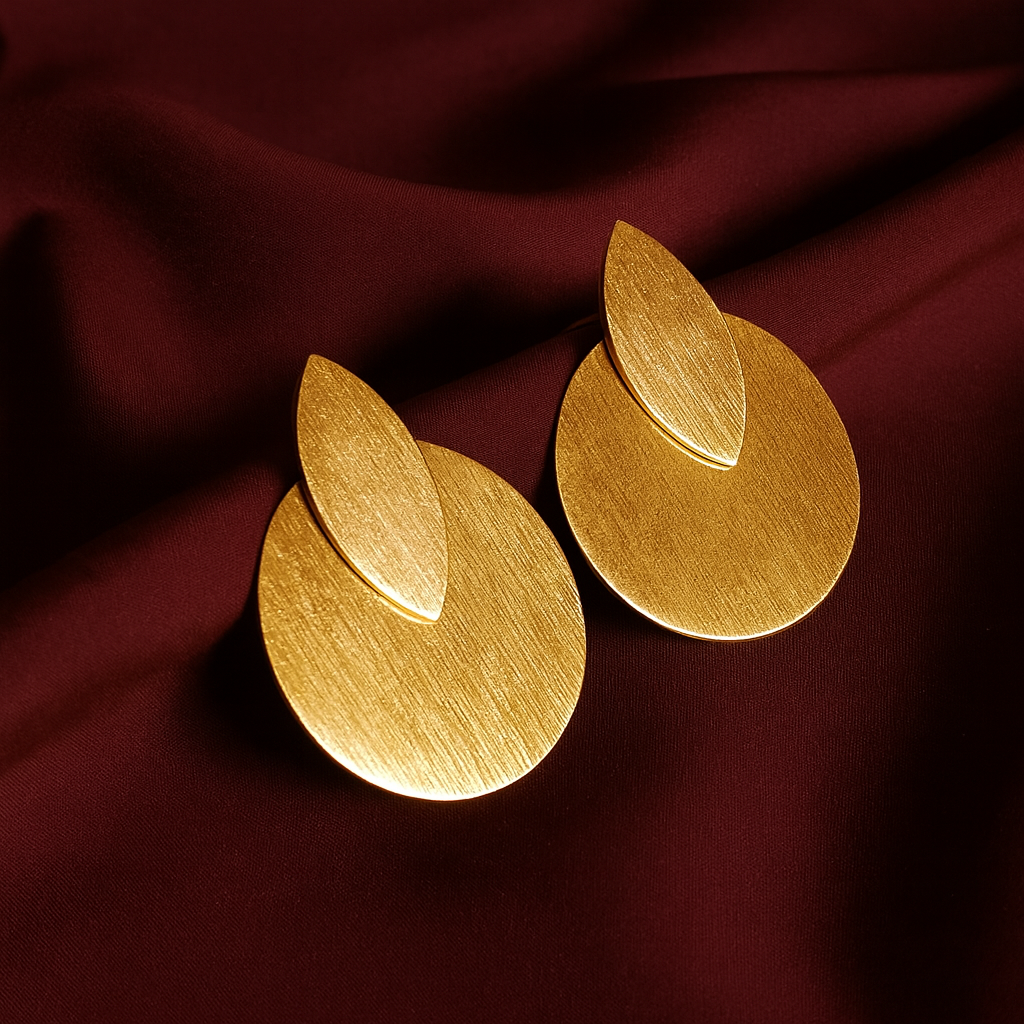 A pair of handcrafted gold-plated earrings featuring a matte brushed finish. The design includes a teardrop-shaped leaf layered over a circular disc, displayed on deep maroon satin fabric. Modern, minimalist, and luxurious.