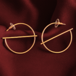 Minimalist gold-plated hoop earrings with a sleek horizontal bar design, Solace earrings by Masaira displayed on maroon satin fabric.