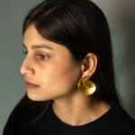 Left profile: model wearing Masaira Sculptural Swirl brushed gold-plated statement stud earring