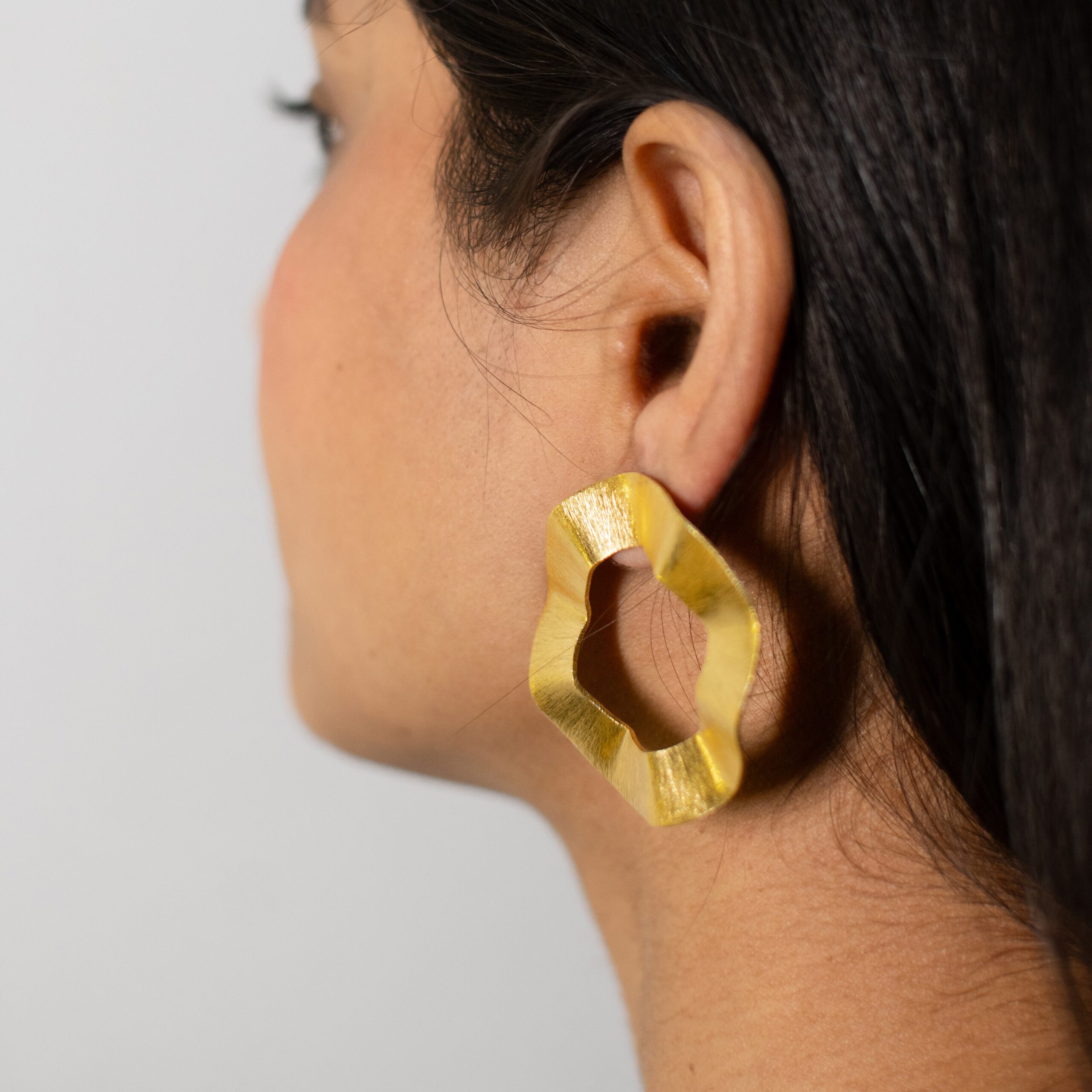 On-ear three-quarter view: model wearing Masaira Ripple Halo brushed gold-plated wavy open-circle stud