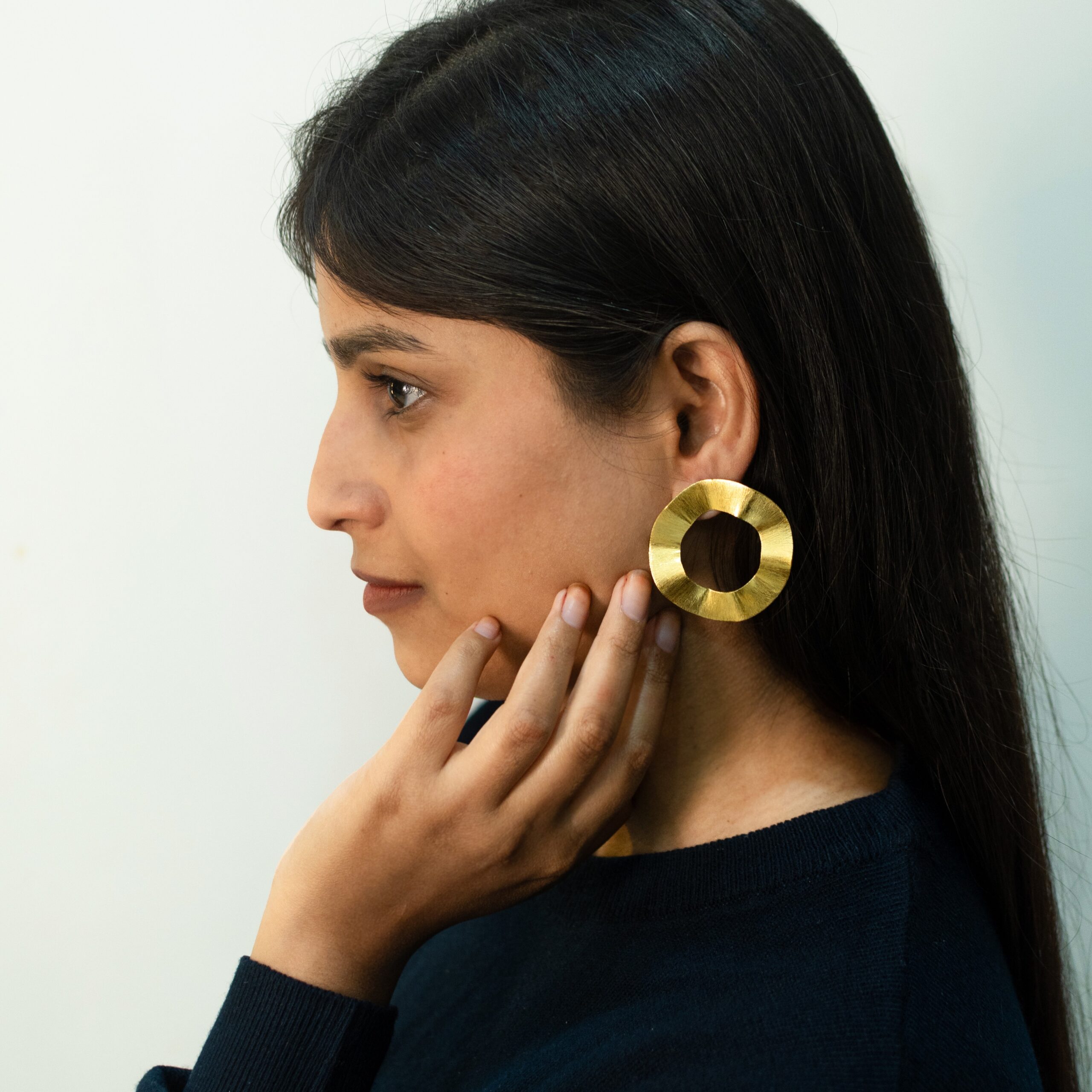 Left profile: model wearing Masaira Ripple Halo brushed gold-plated wavy open-circle statement stud