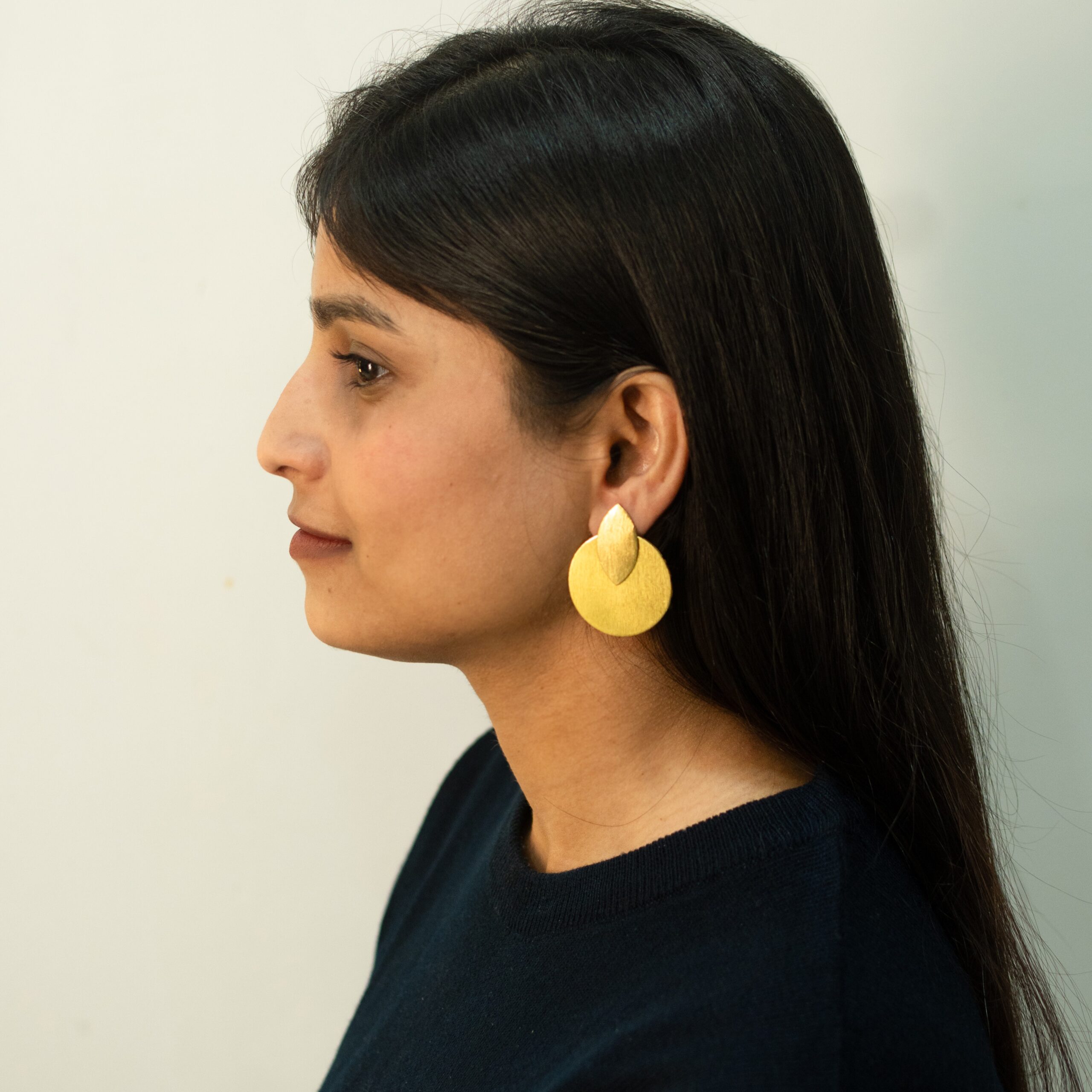 Woman in profile view wearing large matte gold earrings shaped like an overlapping circle and petal.