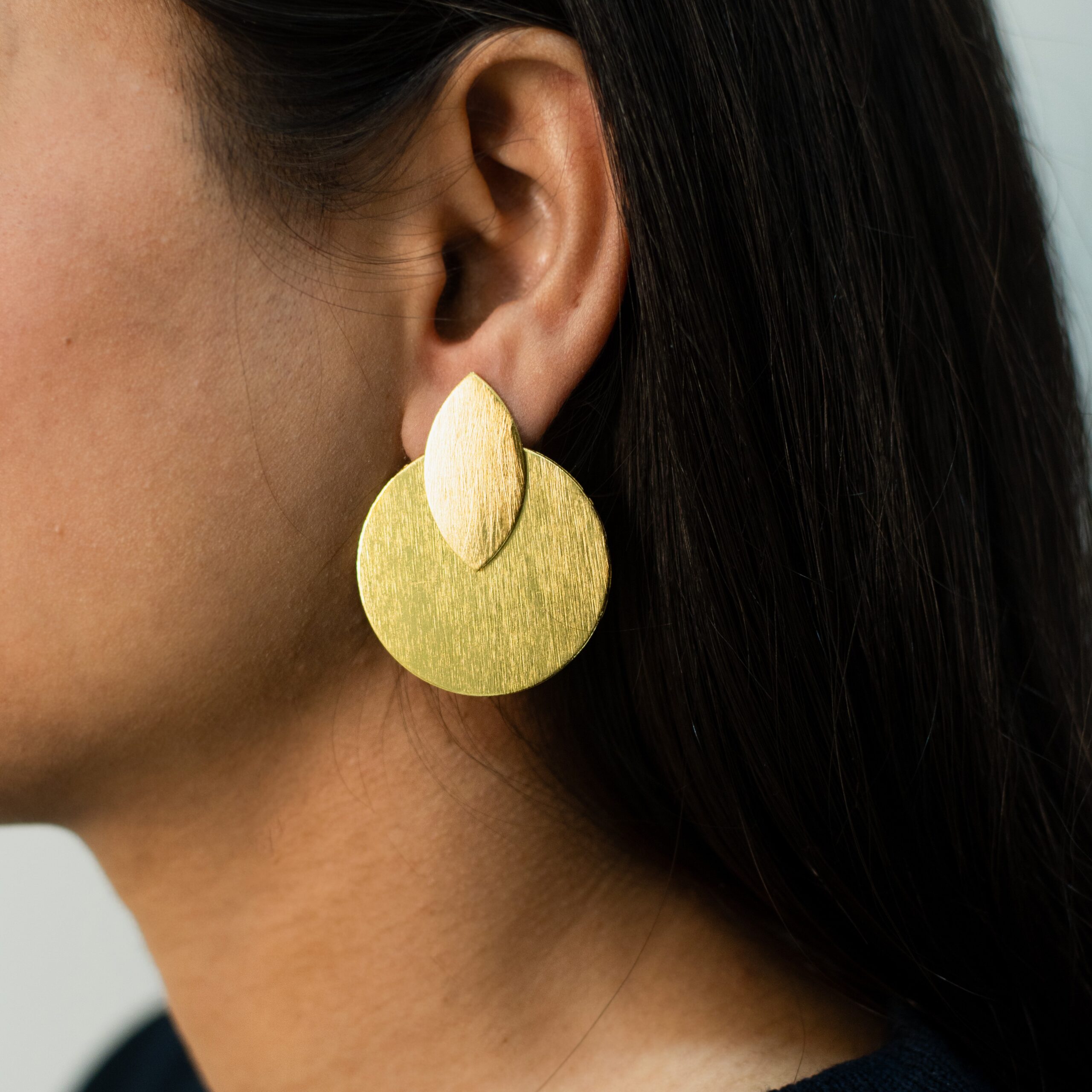 Close-up of a woman wearing matte gold dual-layered geometric earrings with a teardrop and round disc design.