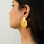 Rear side profile of a woman wearing brushed gold earrings — overlapping circle and teardrop shapes.