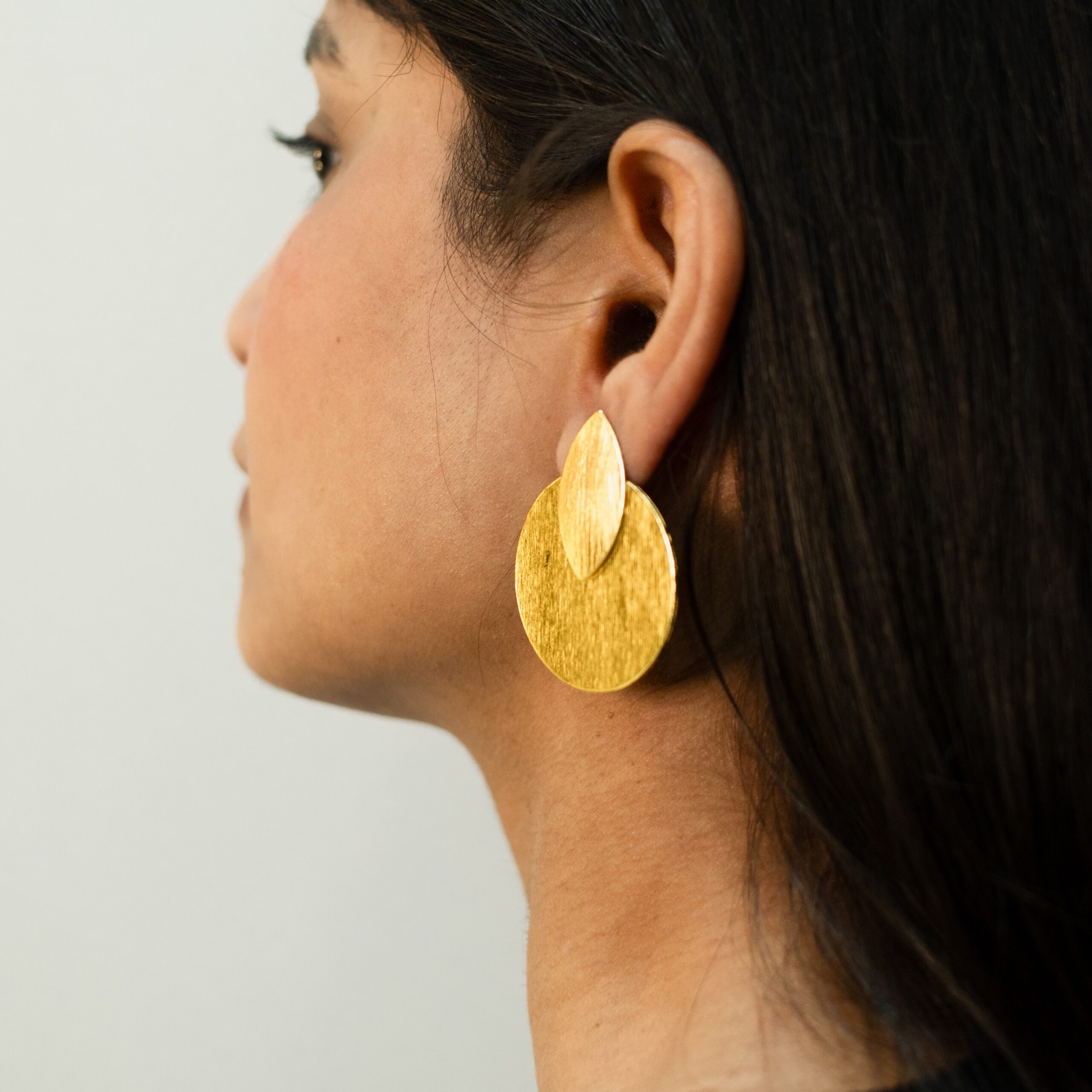 Rear side profile of a woman wearing brushed gold earrings — overlapping circle and teardrop shapes.