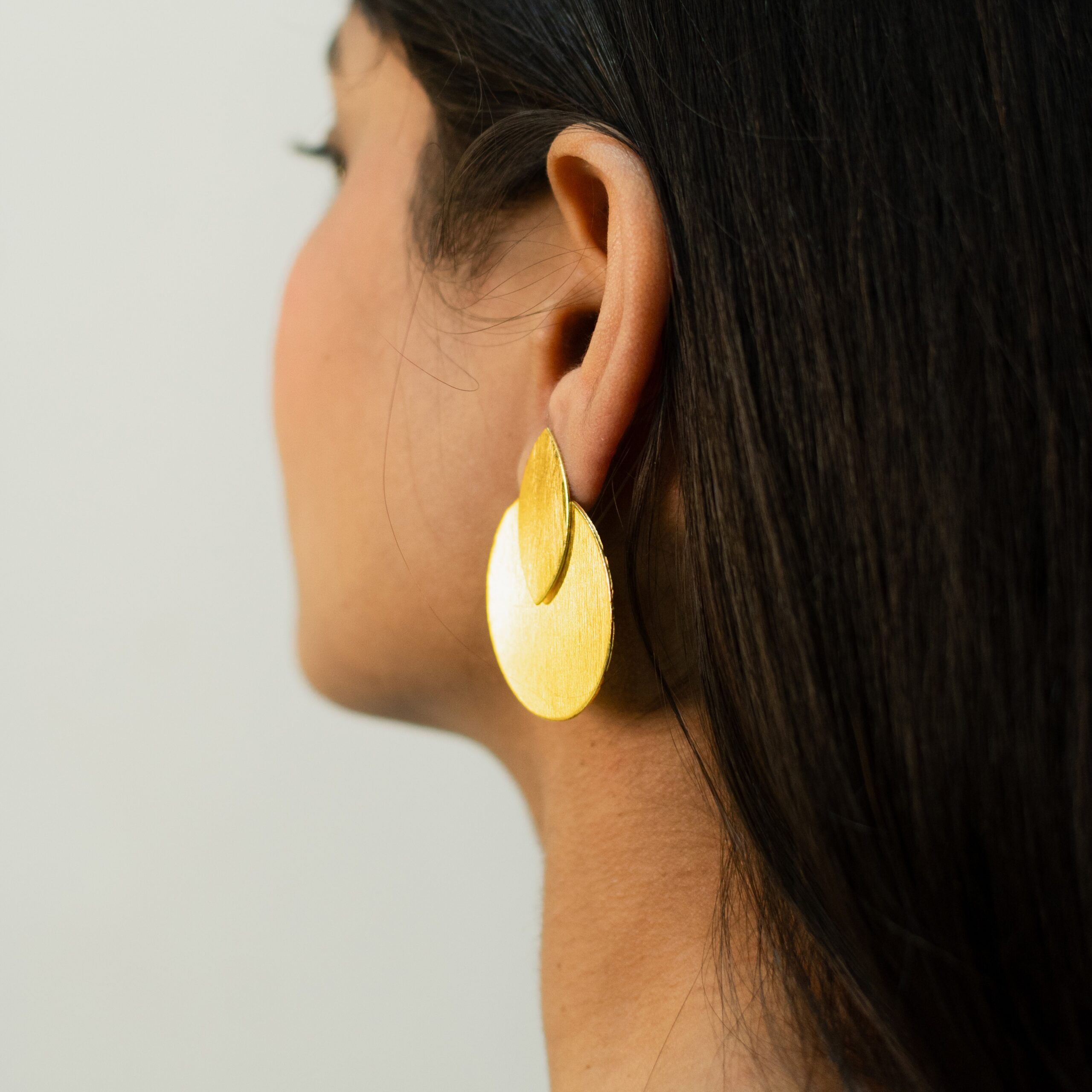 Soft angled close-up of woman’s ear with teardrop-over-disc matte gold statement earring.