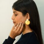 A woman gently touching her chin, showcasing bold matte gold earrings in a teardrop and circle shape.