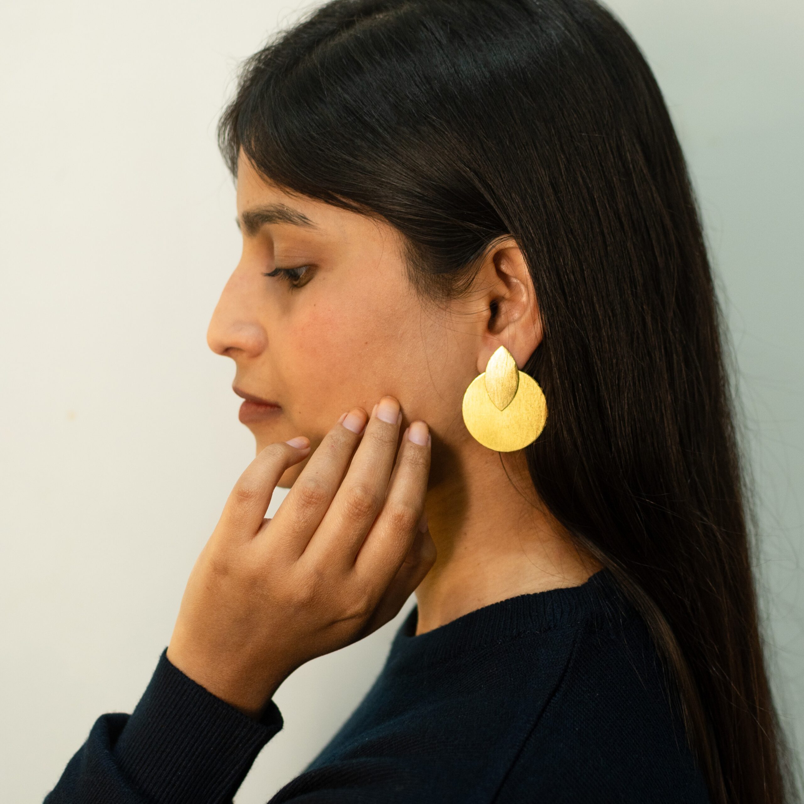 A woman gently touching her chin, showcasing bold matte gold earrings in a teardrop and circle shape.