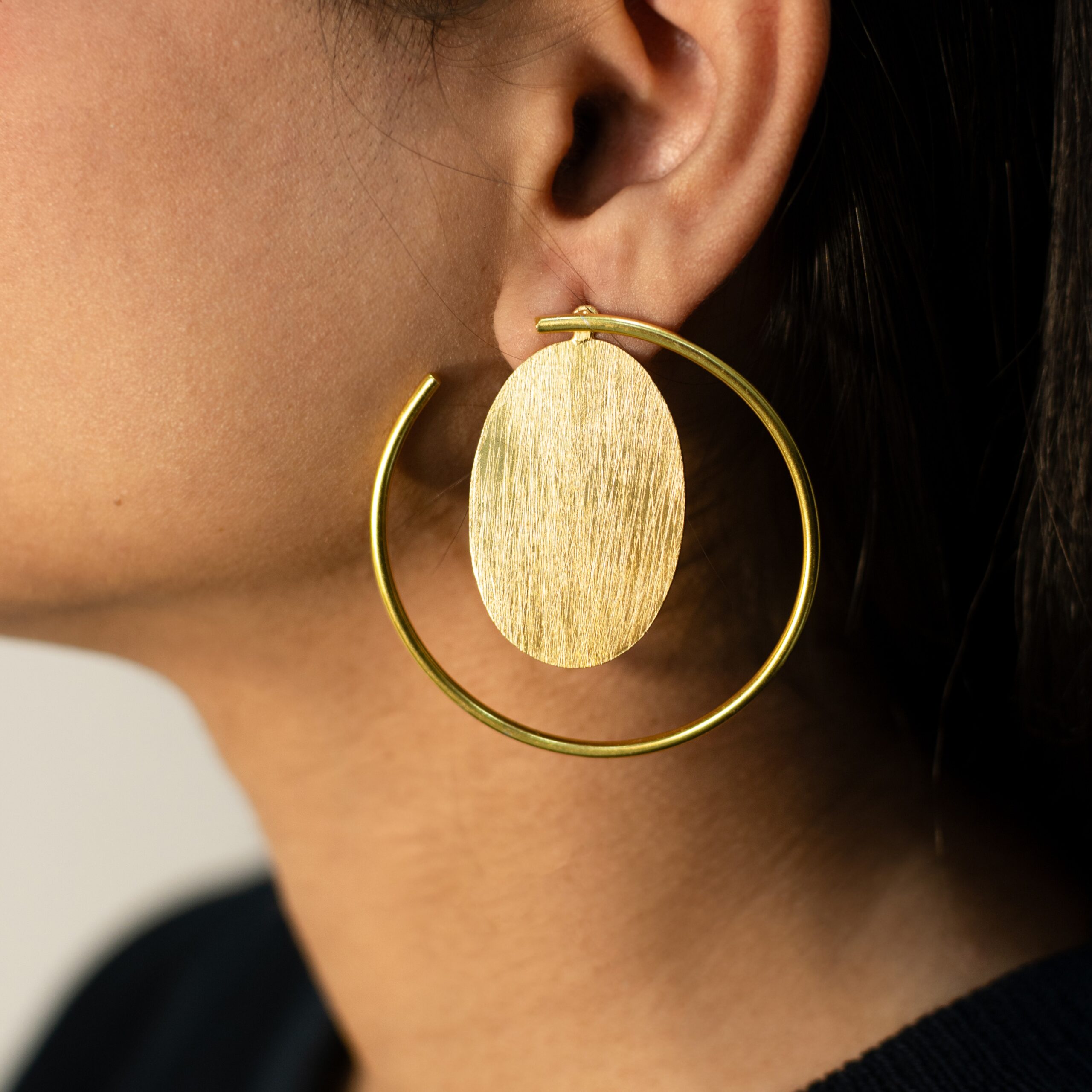 Close-up left ear wearing Masaira Orbit gold-plated open-hoop earring with brushed oval disc