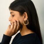 Left profile: model wearing Masaira Double Arc gold-plated open hoop with twin bands, hand on cheek