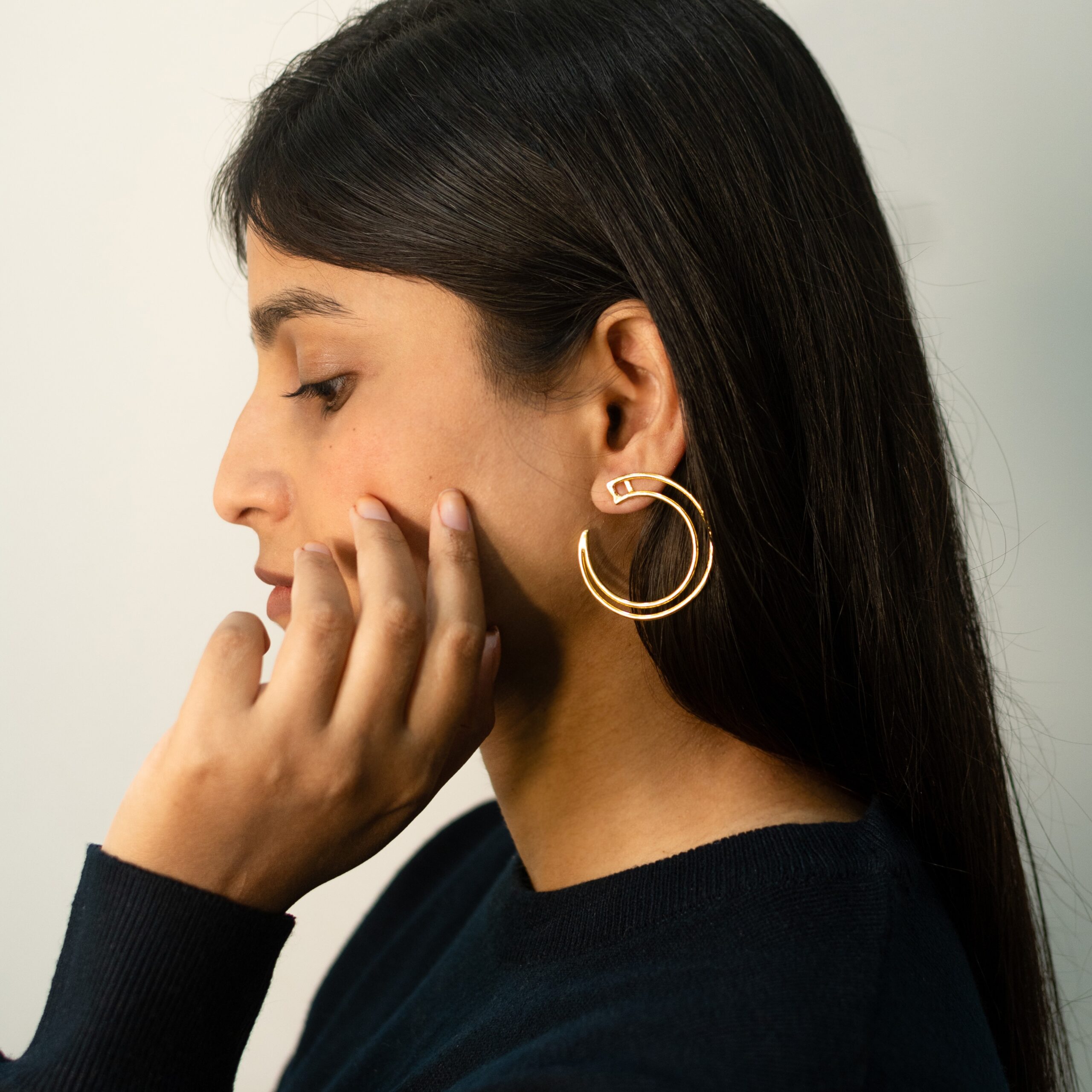 Left profile: model wearing Masaira Double Arc gold-plated open hoop with twin bands, hand on cheek