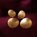 Gold textured double-disc statement earrings with a brushed matte finish on a deep maroon background.