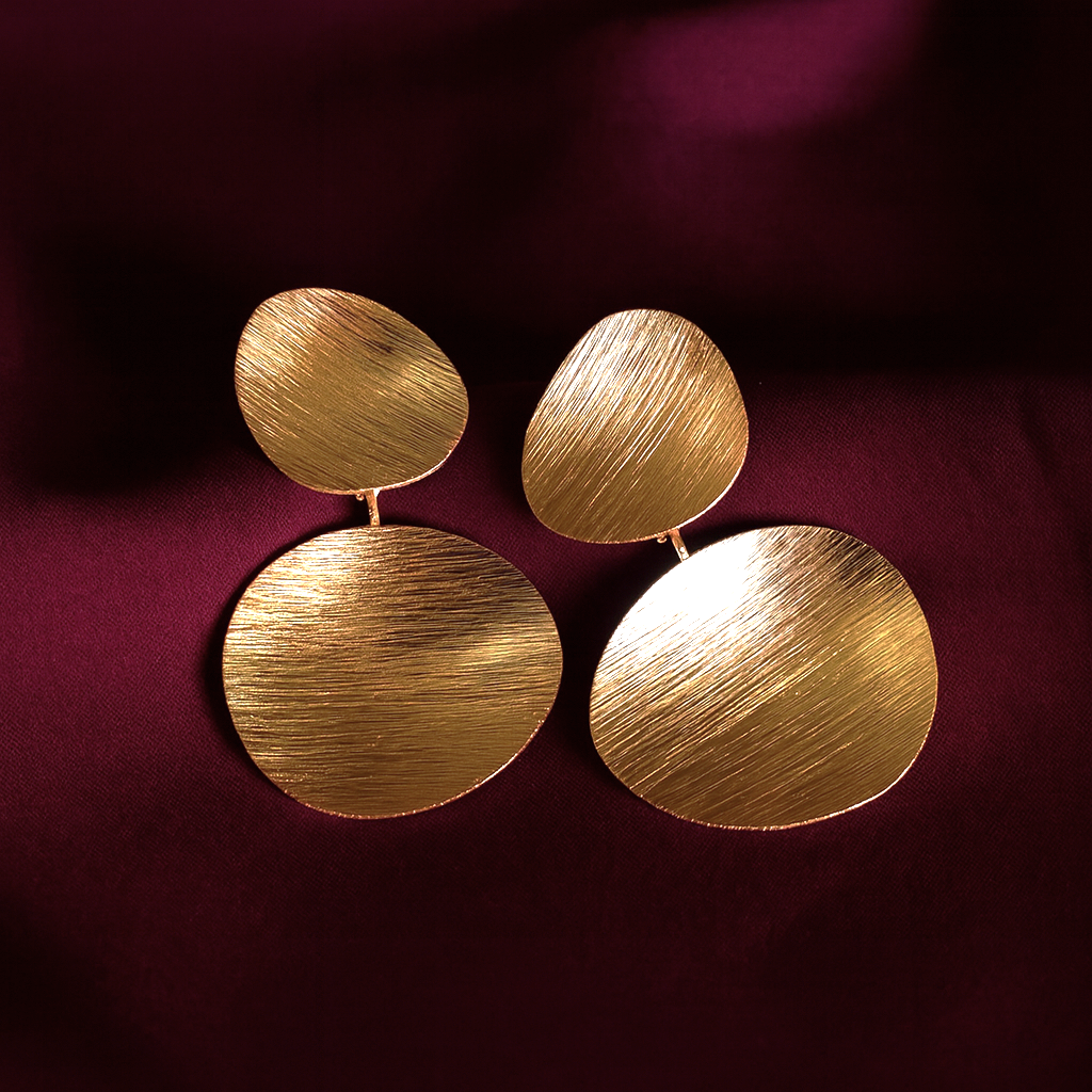 Gold textured double-disc statement earrings with a brushed matte finish on a deep maroon background.