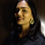 Front-facing portrait of a woman wearing gold cut-out geometric earrings with black outfit against a wooden backdrop.
