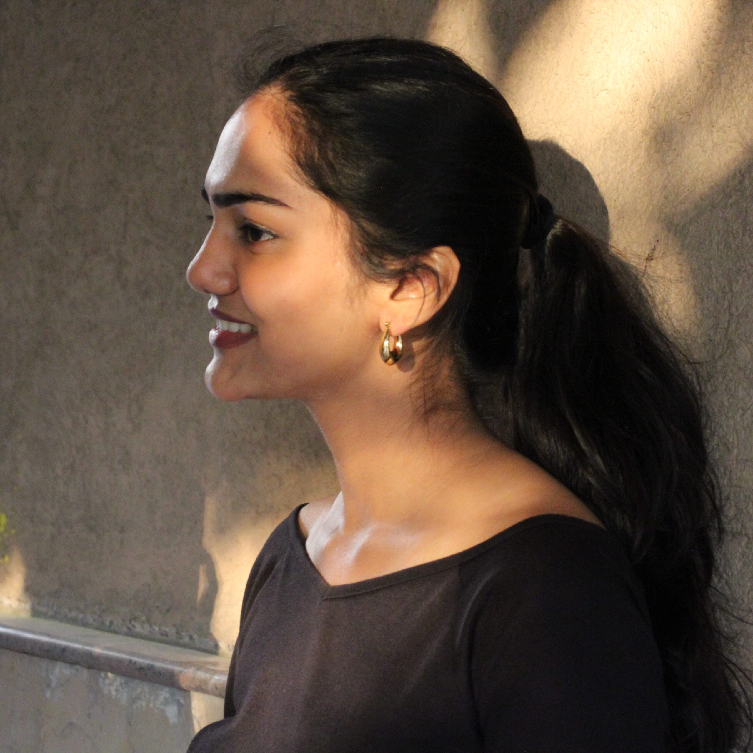 Close side profile of model smiling in Masaira Luna Drop teardrop huggie earrings, glossy gold finish in natural light.