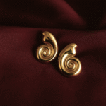 Gold-plated swirl design earrings with a polished spiral finish placed on a maroon satin background. Statement jewelry by Masaira.