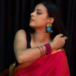Model adjusting hair while wearing Krishna face earrings in red saree — devotional ethnic jewellery by Masaira