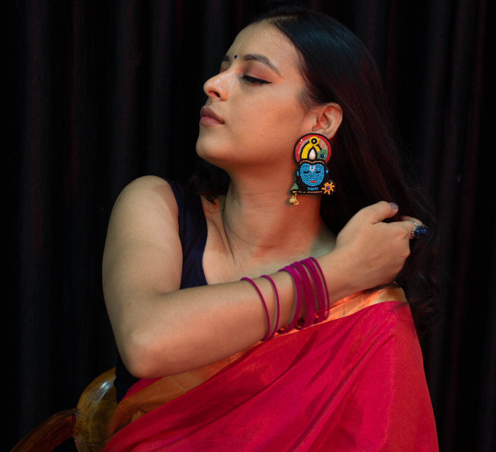 Model adjusting hair while wearing Krishna face earrings in red saree — devotional ethnic jewellery by Masaira