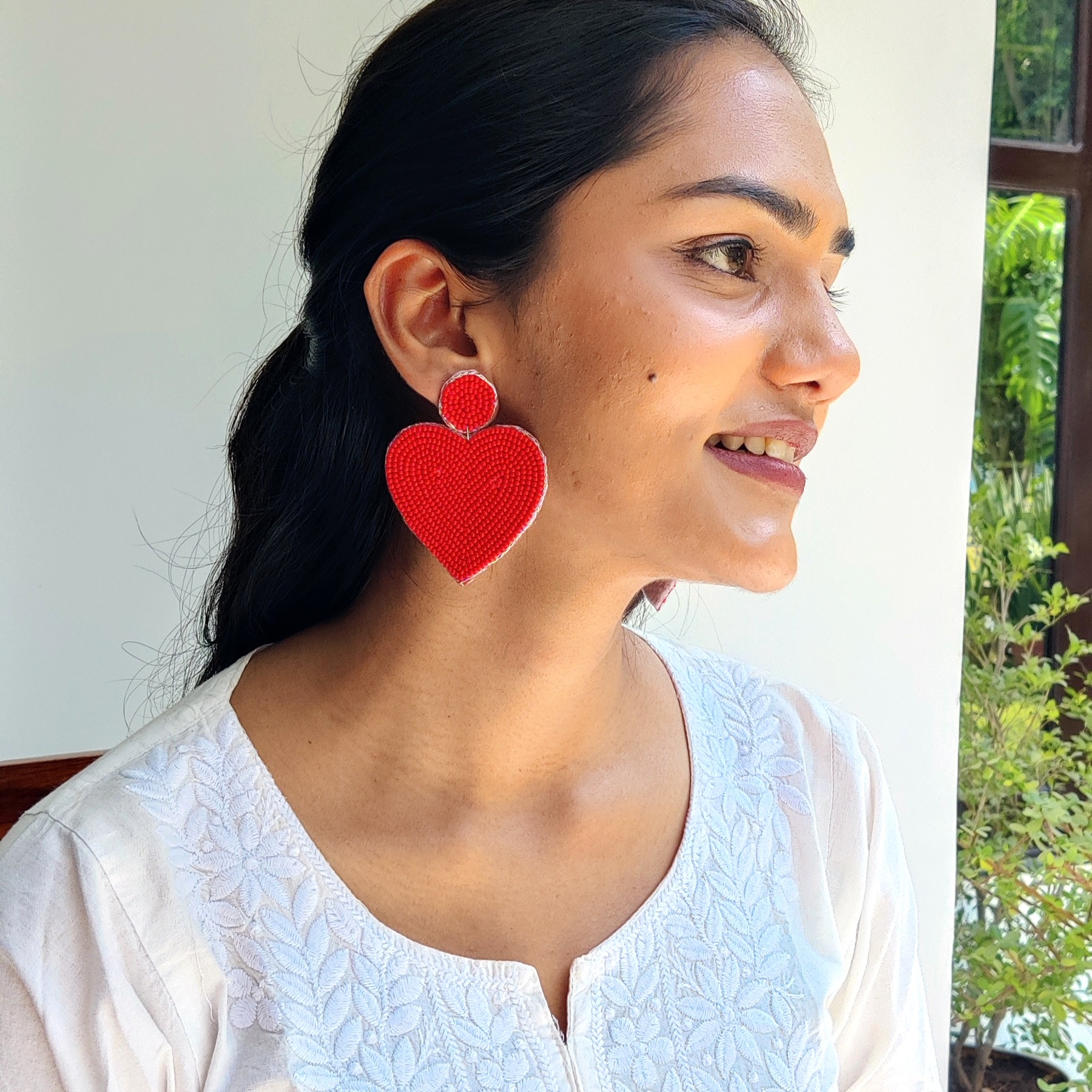 Side-profile lifestyle shot of model by a window wearing red beaded heart earrings — Masaira jewellery, natural light