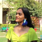 Woman smiling in green dress wearing blue feathered parrot earrings in an urban park.