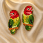 A pair of handcrafted beaded earrings shaped like colorful parrots, featuring red, green, yellow, and orange sequins, placed on a luxurious champagne satin background.