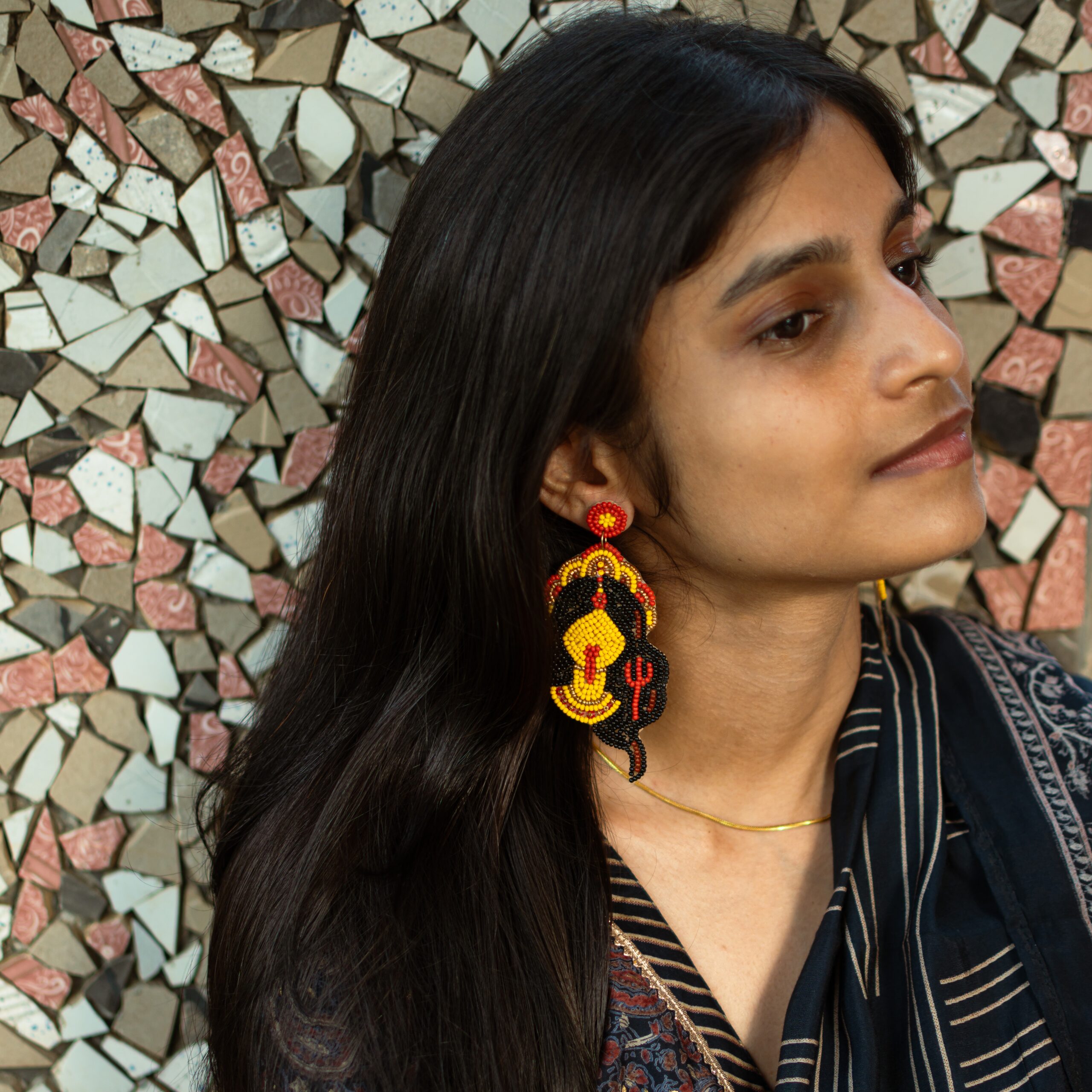 Indian woman wearing handcrafted Kali beaded earrings representing the Hindu goddess Kali, photographed against a mosaic wall.