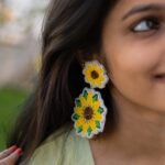 Handcrafted sunflower beaded earrings worn by a smiling woman in a green outfit, outdoor close-up.