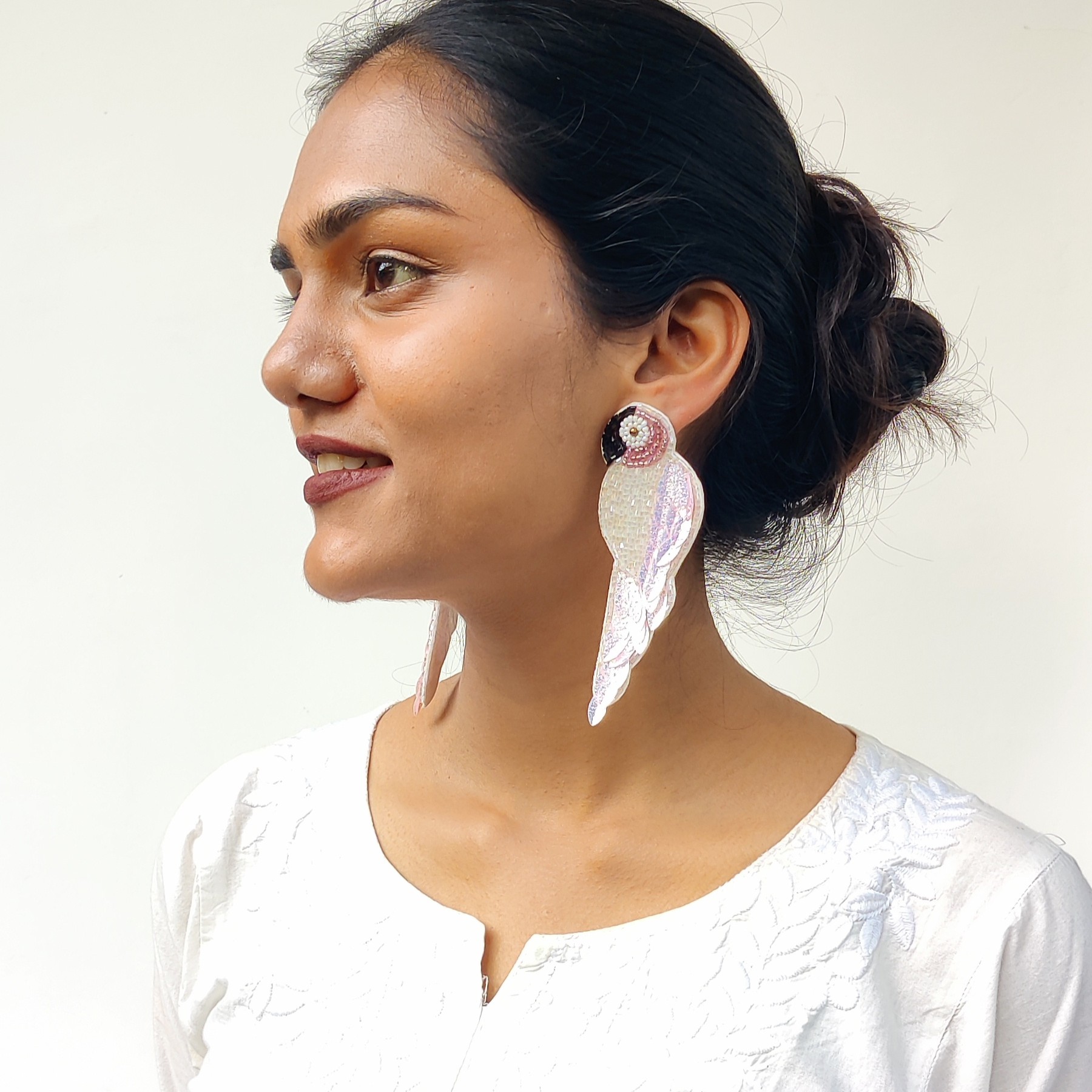 Side profile of woman wearing iridescent pearly parrot beaded earring with a sleek bun and white embroidered top.