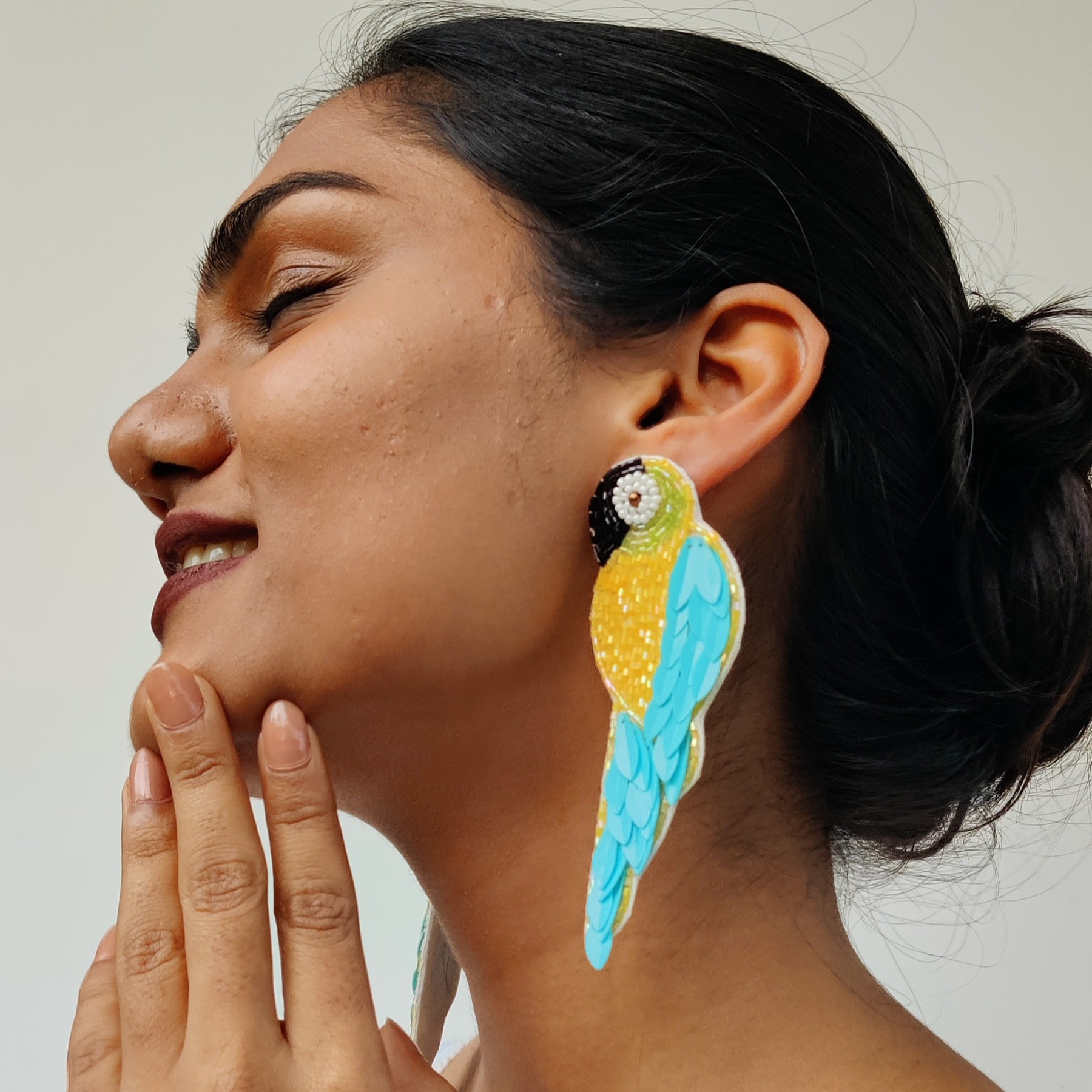 Indian woman smiling while wearing a single large parrot-shaped earring with teal-blue wings and golden beads.