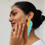 Close-up side profile of a woman wearing a handcrafted teal-blue and golden beaded parrot earring, smiling gently with her hand on her chin.