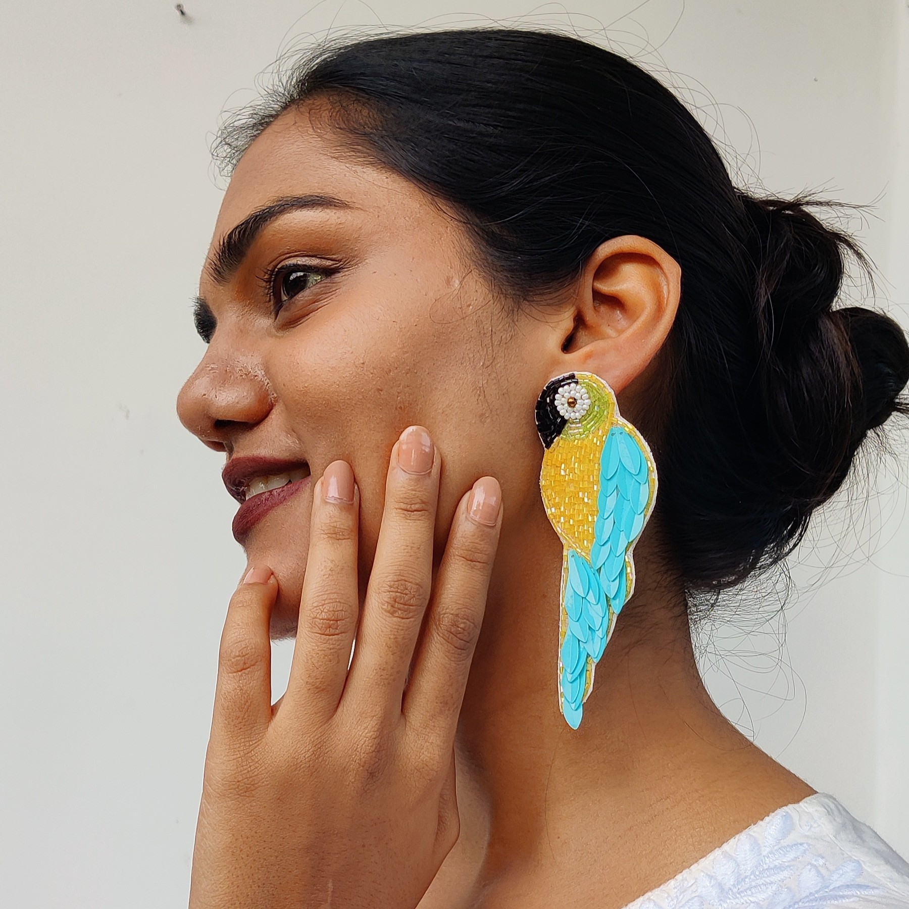 Close-up side profile of a woman wearing a handcrafted teal-blue and golden beaded parrot earring, smiling gently with her hand on her chin.