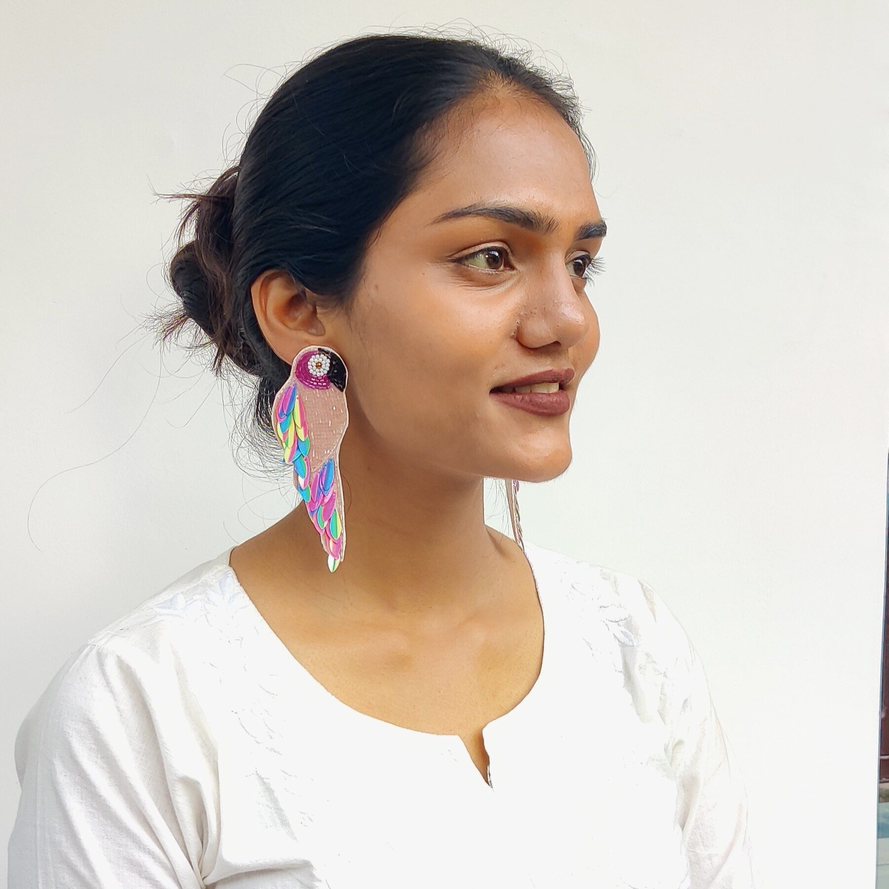 Side-angle lifestyle shot of model in white kurta wearing multicolour parrot earrings with rainbow sequin feathers — Masaira statement jewellery