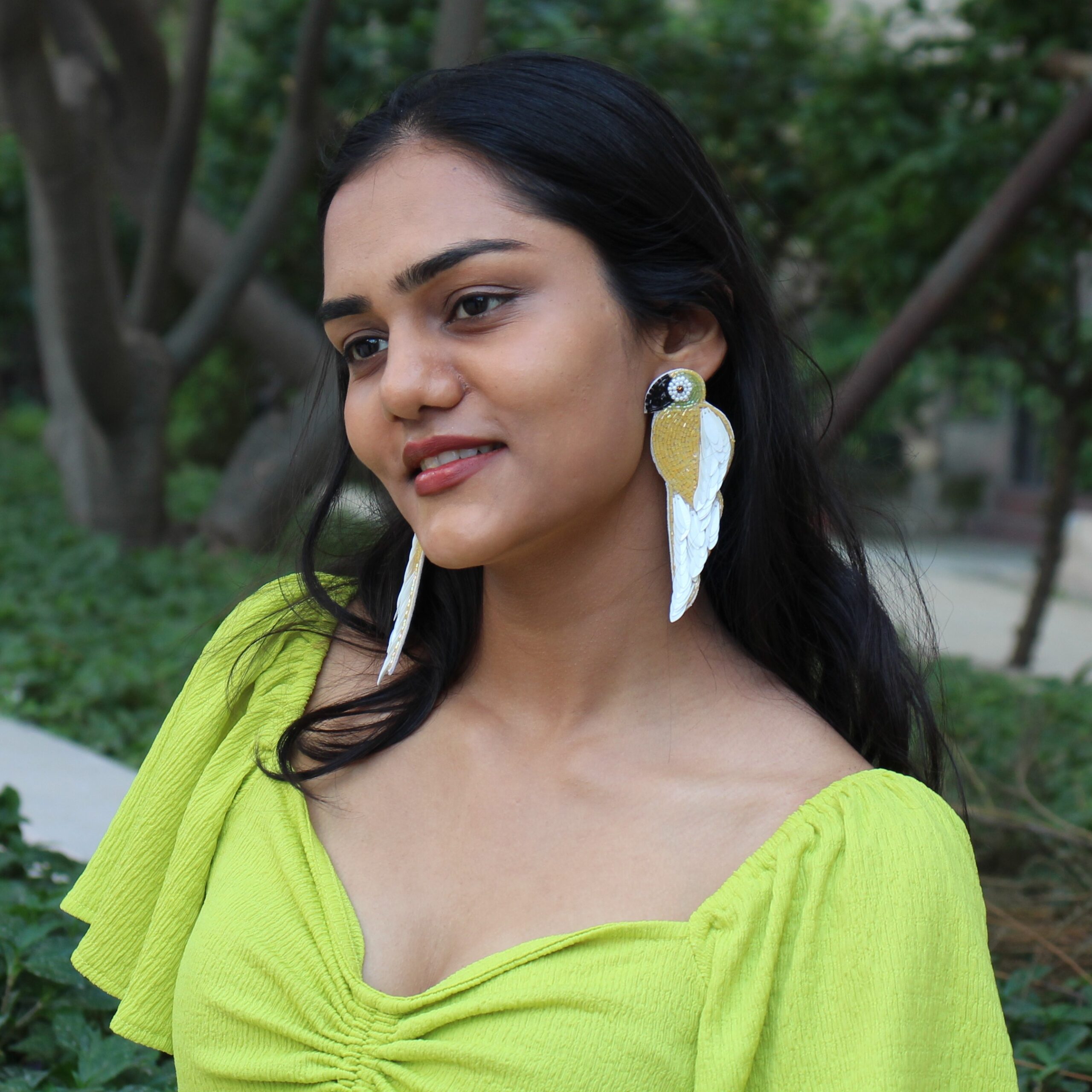 Close-up of woman wearing feather parrot earrings with trees and garden in the background.