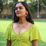 Indian woman in a lime green dress wearing beaded toucan earrings with red flowers and green leaves, standing outdoors on a sunny day.