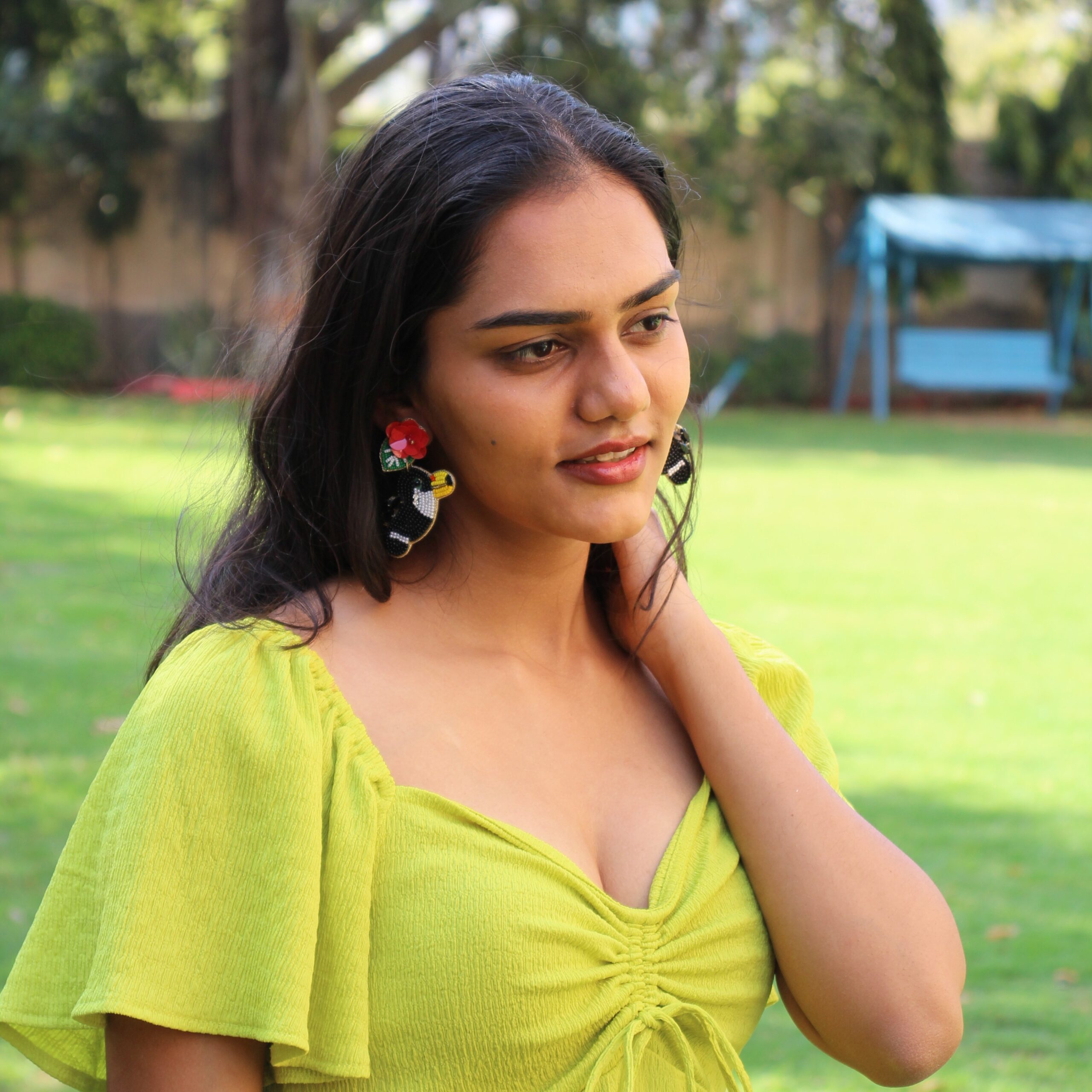 Tropical Toucan Earrings – Handcrafted Jungle Glam Close-up of a woman in green dress wearing large toucan-shaped beaded earrings with red flowers, smiling outdoors.