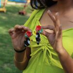 Close-up of a woman’s hands holding toucan-themed beaded earrings with red flower and green leaf details, wearing a lime green outfit.