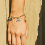 Evil eye gold charm bracelet with blue stone accents, worn on wrist against neutral wall