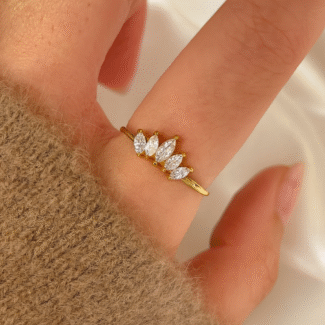 A woman's hand wearing a gold-plated ring with five marquise-cut stones, captured in a close-up shot over an ivory satin background