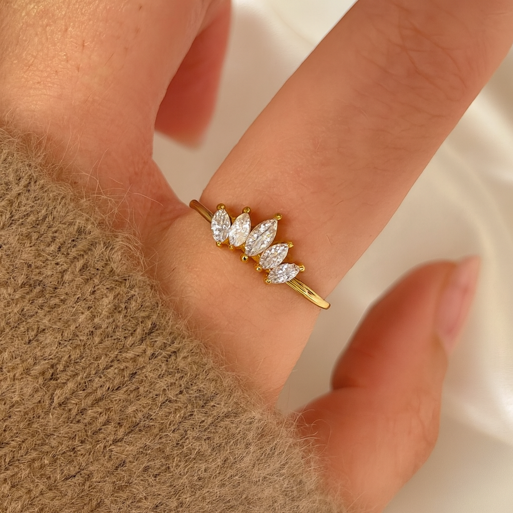 A woman's hand wearing a gold-plated ring with five marquise-cut stones, captured in a close-up shot over an ivory satin background