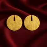 Gold-plated round minimalist earrings with vertical slits, placed on a rich maroon satin fabric background.