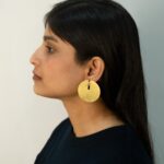 Full side profile of a woman wearing a minimalist round gold earring with a cutout, styled with straight black hair.