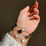 Woman wearing stacked gold-plated black and white clover charm bracelets on wrist in natural daylight