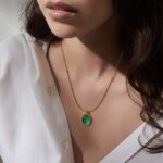 Emerald Aura Necklace with green oval stone pendant on 18 carat gold plated chain, worn with a white blouse, anti-tarnish and hypoallergenic