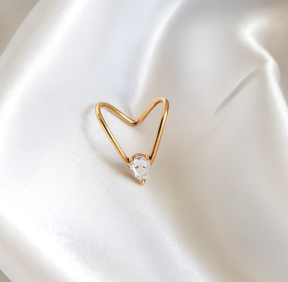 Heart-shaped open gold ring with a clear round stone placed on satin white background